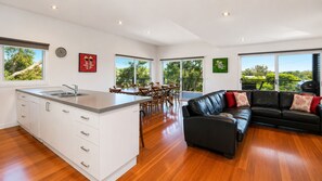 Cocina moderna con una isla central, comedor con sillas, sofá de cuero negro y amplios ventanales con vistas a zonas verdes.