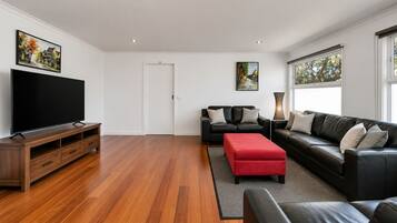 A modern living room with a flat-screen TV, black leather sofas, a red ottoman, and hardwood flooring.
