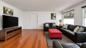 A modern living room with a flat-screen TV, black leather sofas, a red ottoman, and hardwood flooring. - Rumah Pantai by Ready Set Stay- Unlimited WiFi & Close to the Beach (Smiths Beach)
