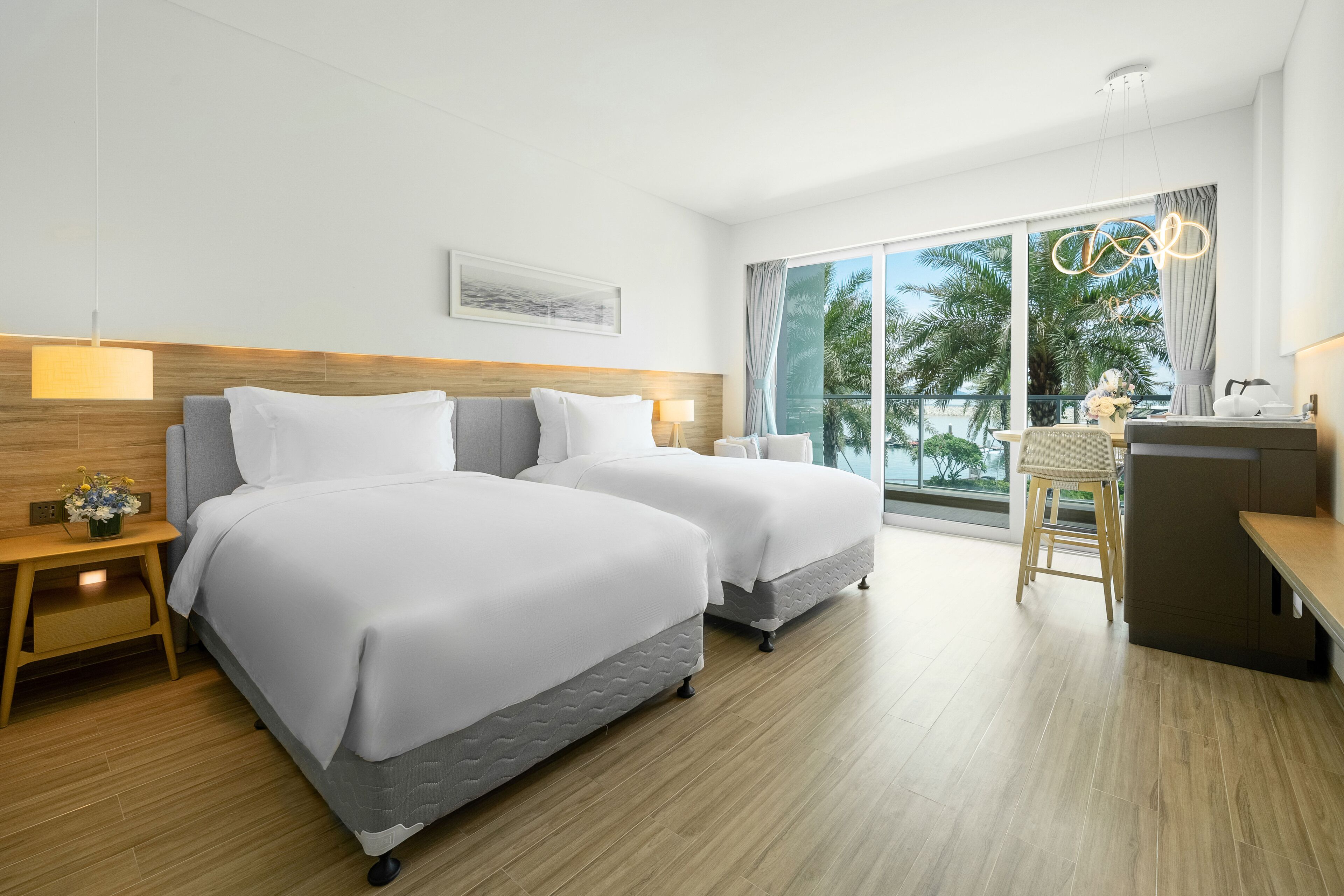 A hotel room with two beds, a desk, and a balcony with a view of palm trees.
