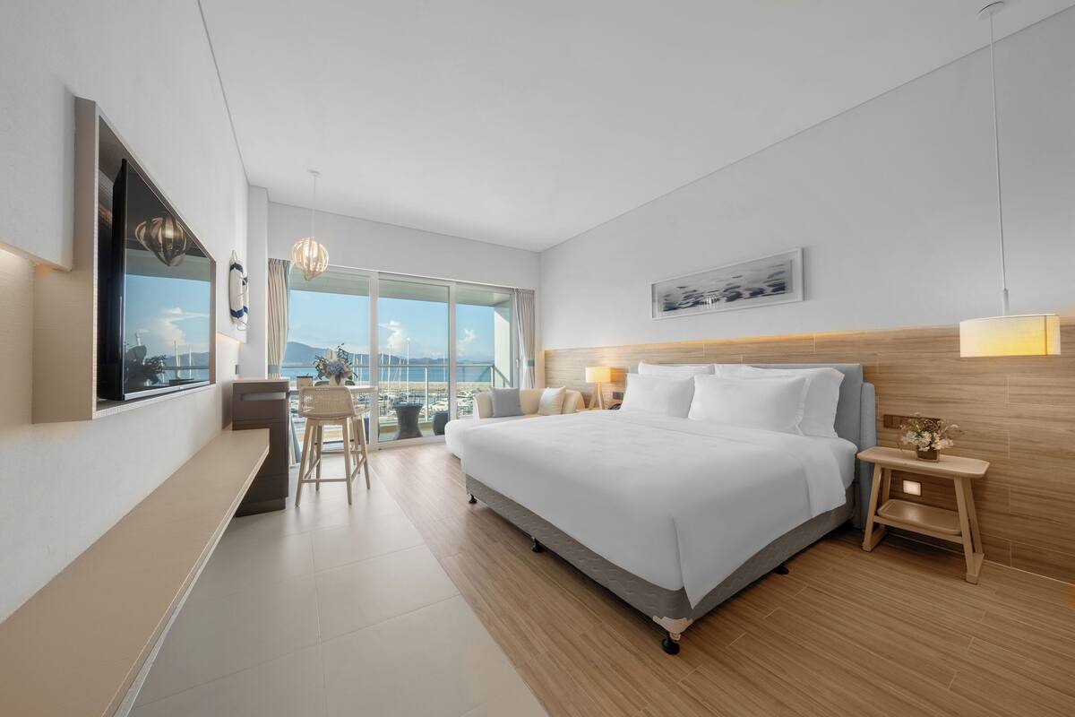 Habitación de hotel moderna con una cama grande, un escritorio, una silla, una mesita de noche con lámpara y vistas a la ciudad a través de amplias ventanas.
