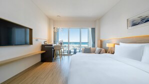 Ein Hotelzimmer mit einem großen Bett, einem Schreibtisch mit Stuhl, einem an der Wand befestigten Fernseher und Meerblick durch große Fenster.