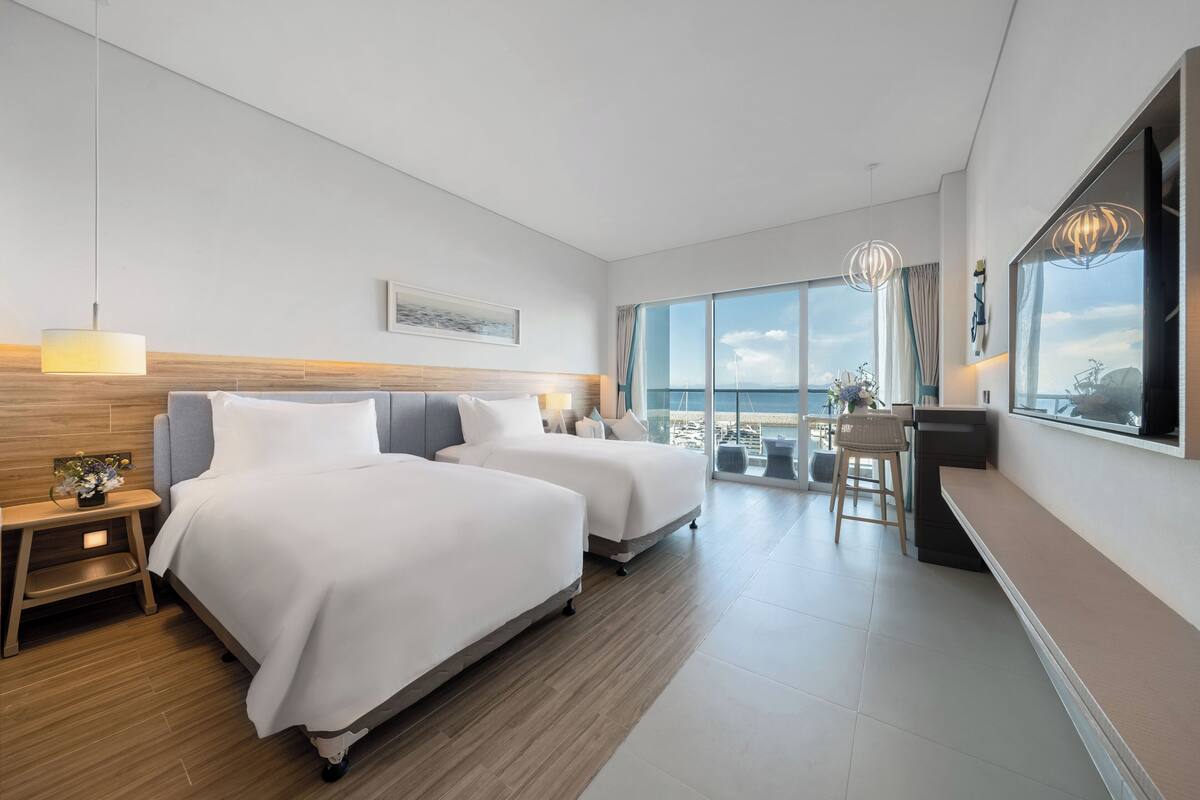 Habitación de hotel con dos camas, un escritorio, una silla y un amplio ventanal con vistas al mar.