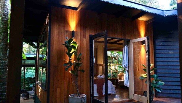 A wooden exterior with a glass door leading to an interior with a bed and a chair. - Ananda Eco House - Rainforest Retreat (Montville)