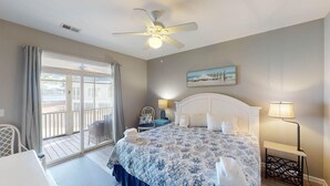 A bedroom with a bed, a ceiling fan, a painting, and a view of the outdoors.