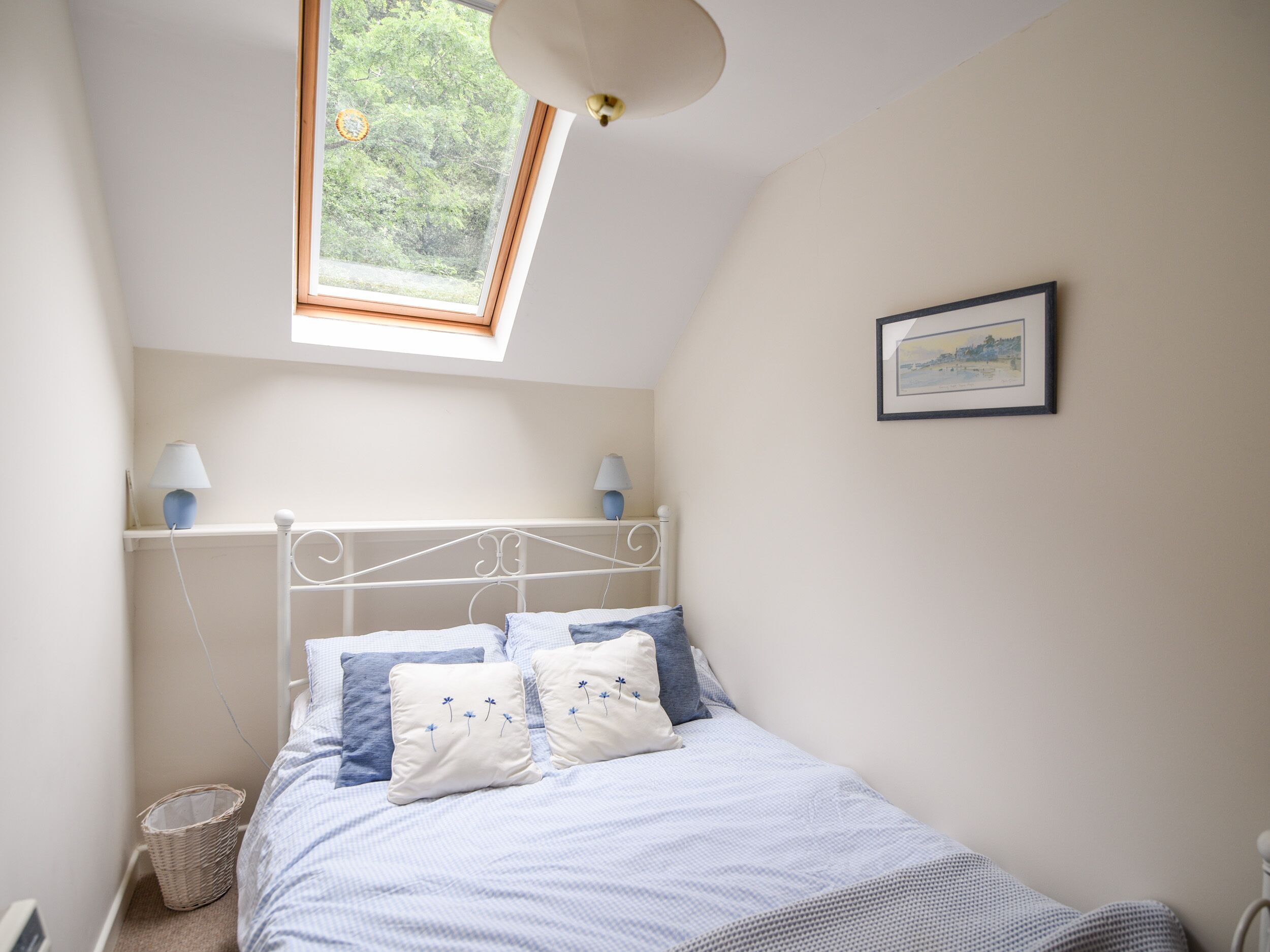Une chambre avec une lucarne, un lit avec une literie bleue et blanche, deux lampes de chevet et un tableau encadré au mur.