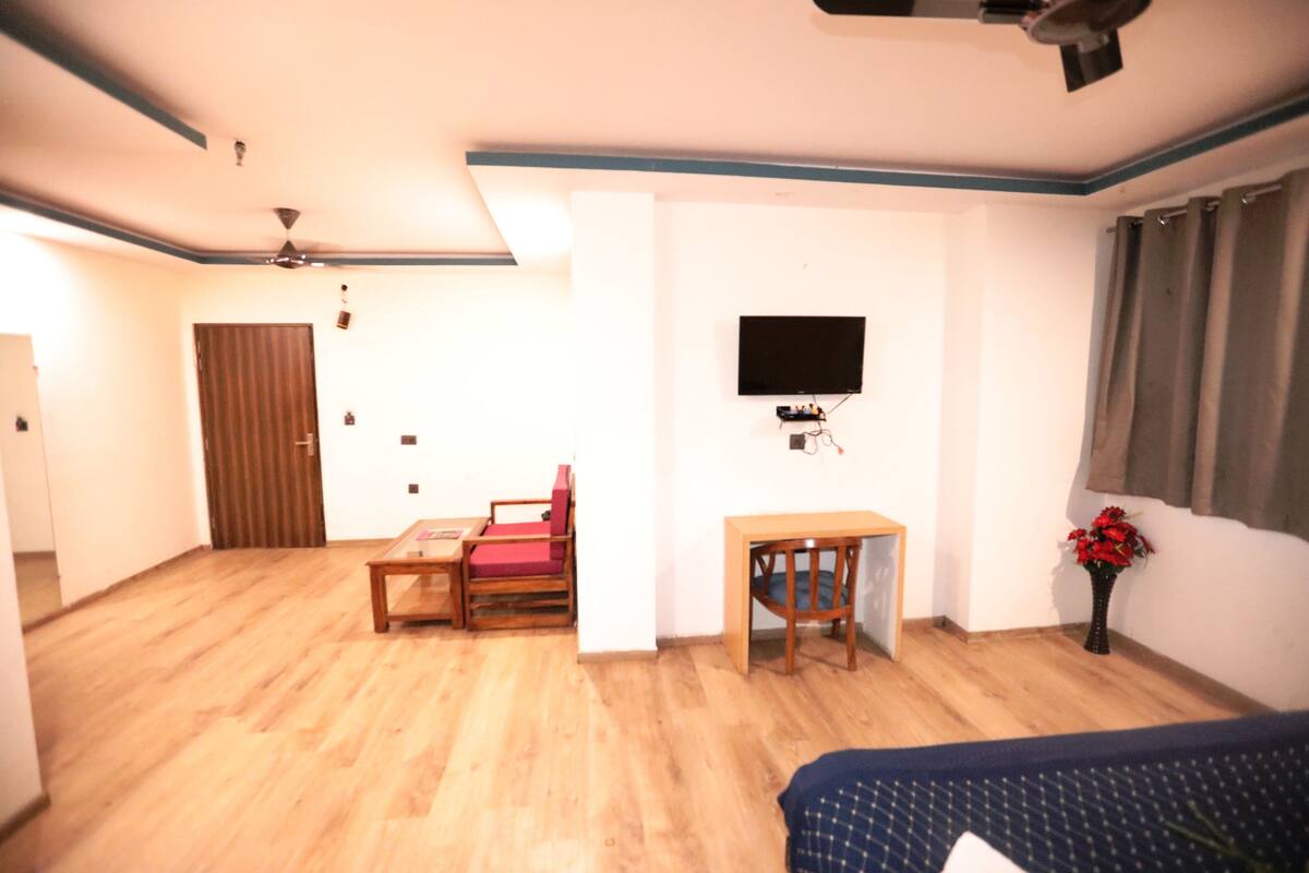 A room with a wooden floor, a bed, a desk, a chair, a television mounted on the wall, and a door.