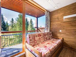A wooden cabin with a balcony, a sofa with a patterned cover, and a view of trees. - Bright renovated studio · Sleeps 4 · South/west balcony (Les Arcs 1800)