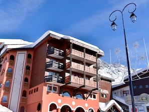 Un edificio de varios pisos con balcones, una pista de esquí al fondo y un letrero que indica un punto de alquiler de esquíes.