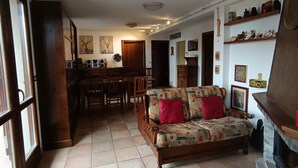 A living room with a sofa, a dining table, and chairs. There is a kitchen area with cabinets and a refrigerator.
