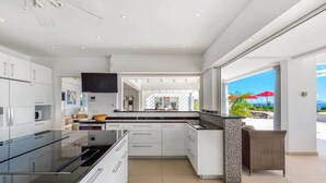 Eine moderne Küche mit weißen Schränken, einer schwarzen Arbeitsplatte und einer großen Kochinsel. Es gibt einen Essbereich mit Korbmöbeln und Blick auf das Meer.