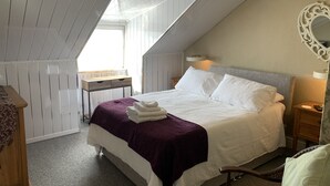 A bedroom with a sloped ceiling, a bed with white linens and a purple throw, a bedside table with a lamp, and a mirror on the wall.