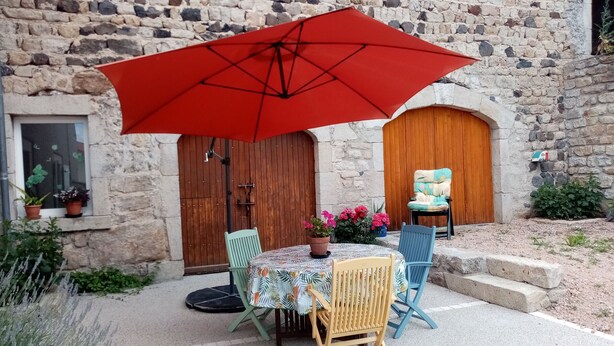 Une terrasse extérieure avec une table et des chaises, un parasol rouge, et un bâtiment en pierre doté de portes et de fenêtres en bois.