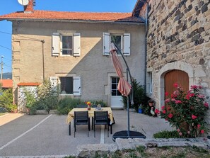 Un bâtiment en pierre avec un toit en tuiles, une table entourée de chaises et un jardin agrémenté de fleurs et de plantes.