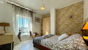 Une chambre à coucher comprenant un lit, une table de chevet, une lampe, une fenêtre avec des rideaux, une armoire et un mur décoré d’un motif.