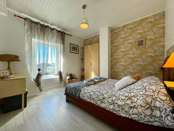Une chambre à coucher comprenant un lit, une table de chevet, une lampe, une fenêtre avec des rideaux, une armoire et un mur décoré d’un motif.
