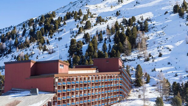 Hotel amb edifici vermell i façana blava, envoltat de muntanyes i arbres coberts de neu.