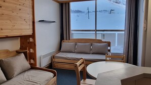 A cozy room with a wooden bunk bed, a small sofa, a round table, and a large window offering a view of a snowy landscape.