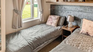 A compact bedroom with two beds, a bedside table with a lamp, and a window with curtains. - 1 bedroom accommodation in Laurenkirk (Laurenkirk)