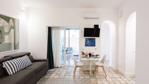 A modern living room with a sofa, a dining table with chairs, a TV, and a view through a glass door to an outdoor area. - Suite Saint Michael (Capri)