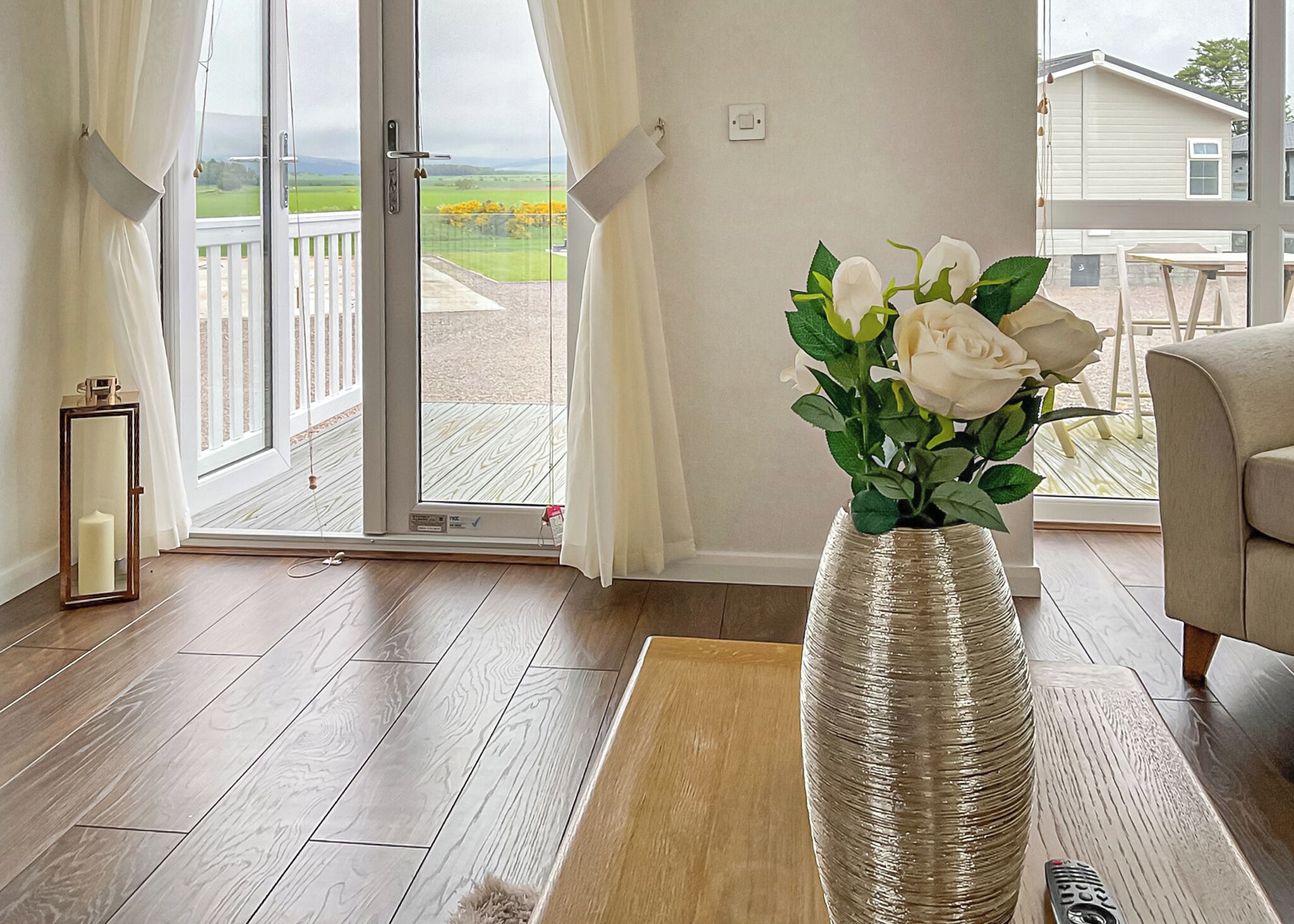 Une pièce avec un plancher en bois, un vase blanc contenant des fleurs blanches, une table d’appoint en bois, un canapé et, à travers une porte vitrée, une vue sur un balcon.
