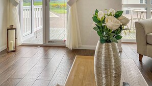 Una habitación con piso de madera, un jarrón blanco con flores blancas, una mesita de madera, un sofá y vistas a un balcón a través de un ventanal.