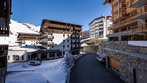 A snow-covered street with traditional wooden buildings, a stone wall, and a parked vehicle.