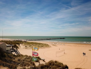 A beach with a wooden pier extending into the sea, a sandy area with flags, and a building with a staircase.