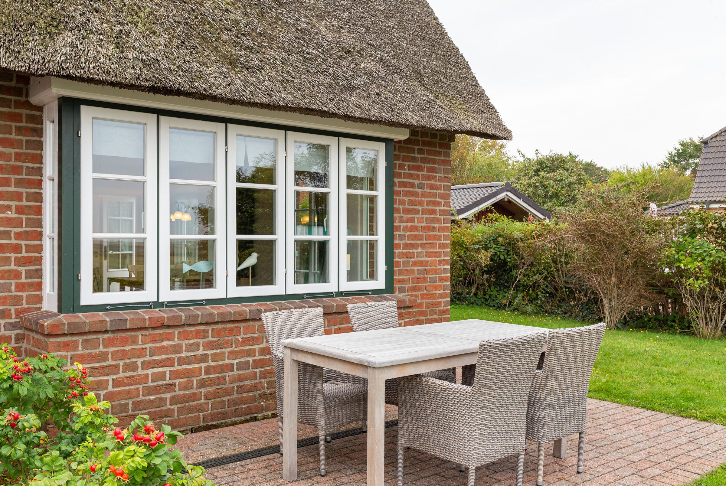 Une maison en briques avec un toit de chaume, une fenêtre à cadre blanc et une terrasse avec une table et des chaises.
