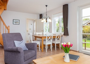 Una sala de estar con un sillón gris, una mesa de comedor de madera, sillas blancas, un jarrón con tulipanes y vistas al jardín a través de un ventanal grande.