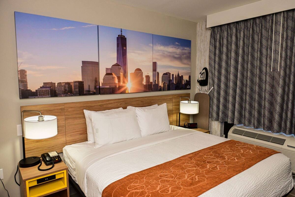 Hotelkamer met een groot stadsgezichtschilderij, een bed met wit bedgoed en een bruine plaid, twee bedlampen, een nachtkastje met een telefoon en een wandairconditioner.