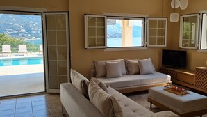 Un saló amb sofà, otomana i televisió. Hi ha finestres amb vistes a una piscina i muntanyes.