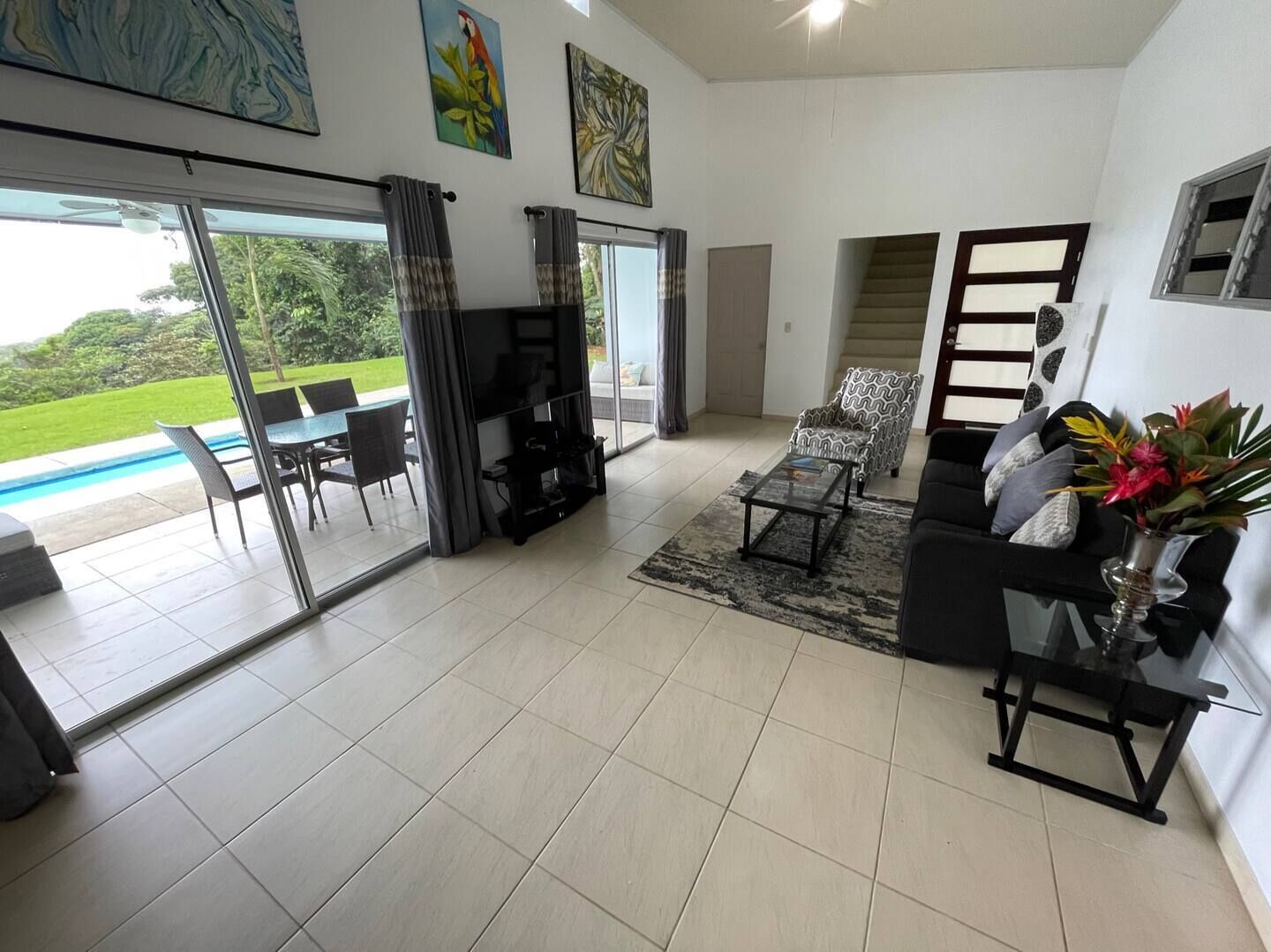 Een ruime woonkamer met een grote glazen schuifpui naar een zwembad, een zwarte bank, een gestreept fauteuil en een flatscreen televisie.