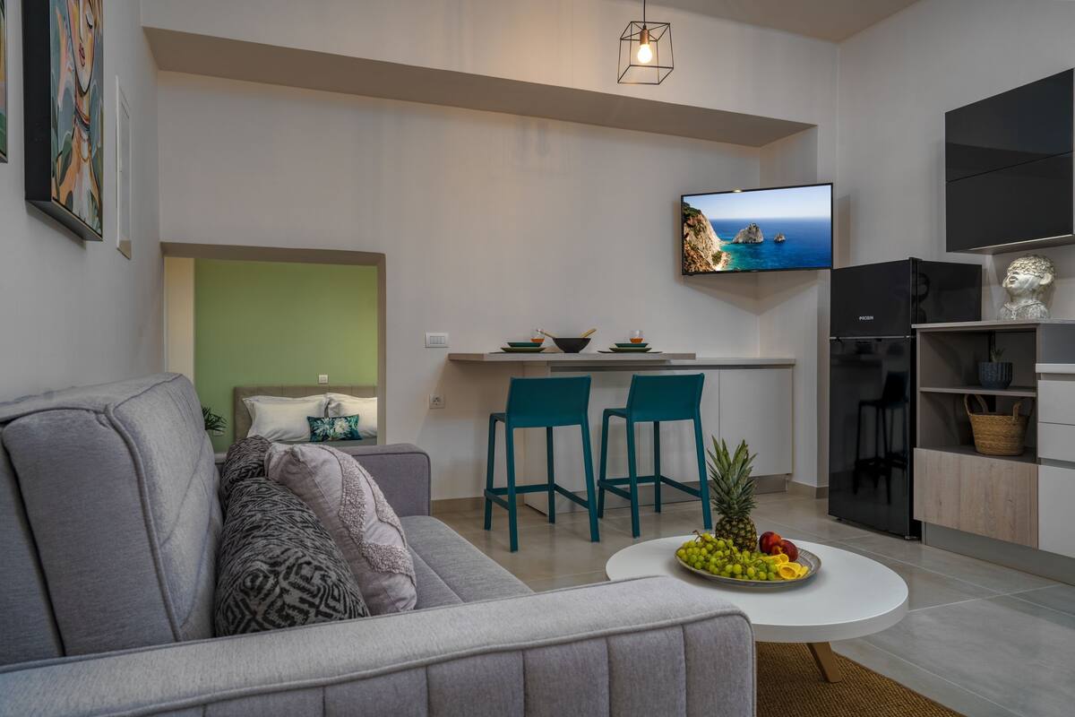 Un salon moderne avec un canapé gris, une table basse sur laquelle on a posé des fruits, des fauteuils de couleur turquoise, un coin cuisine avec un téléviseur et un réfrigérateur.