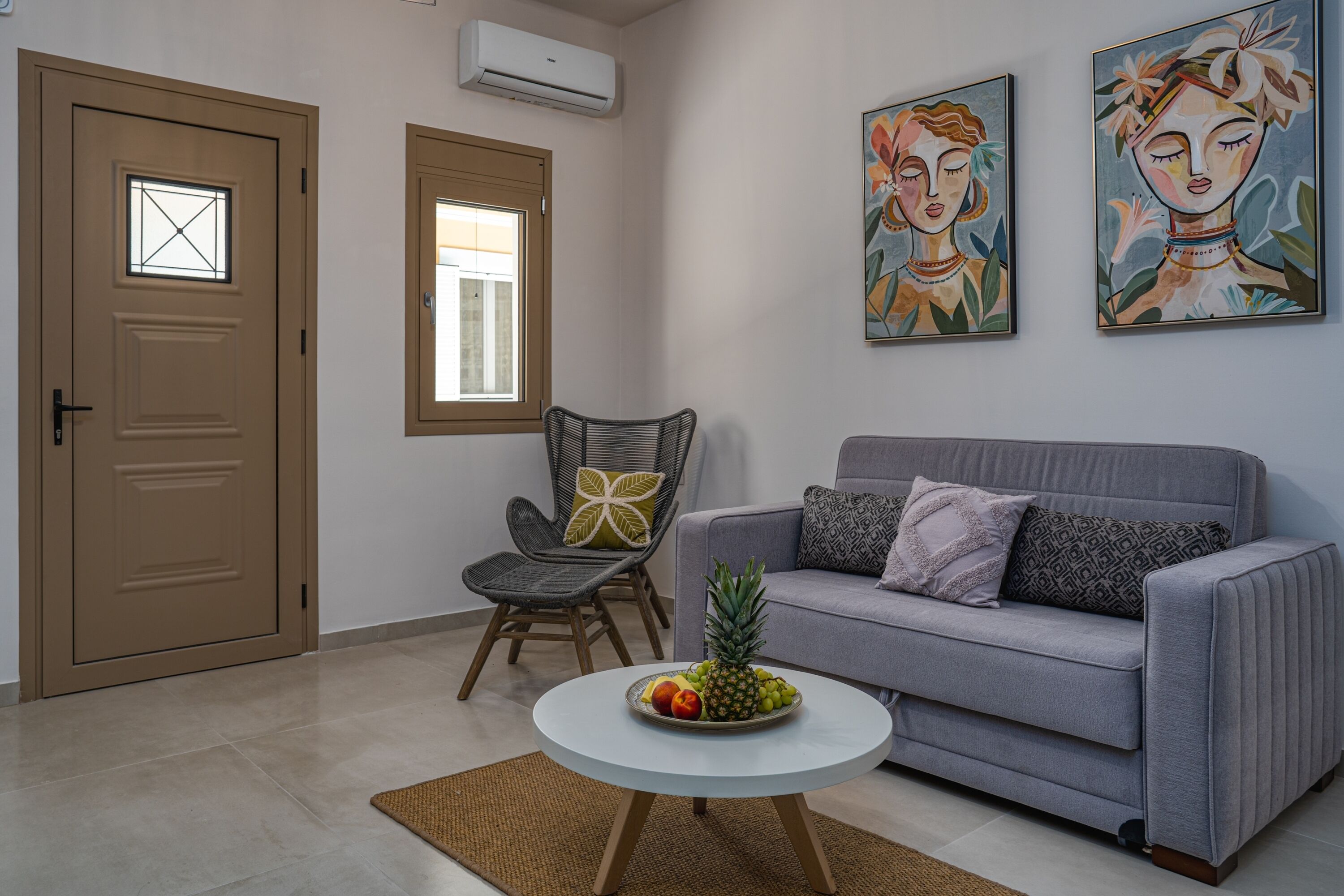 A living room with a grey sofa, a wicker chair, a round coffee table with fruit, and two wall-mounted paintings.