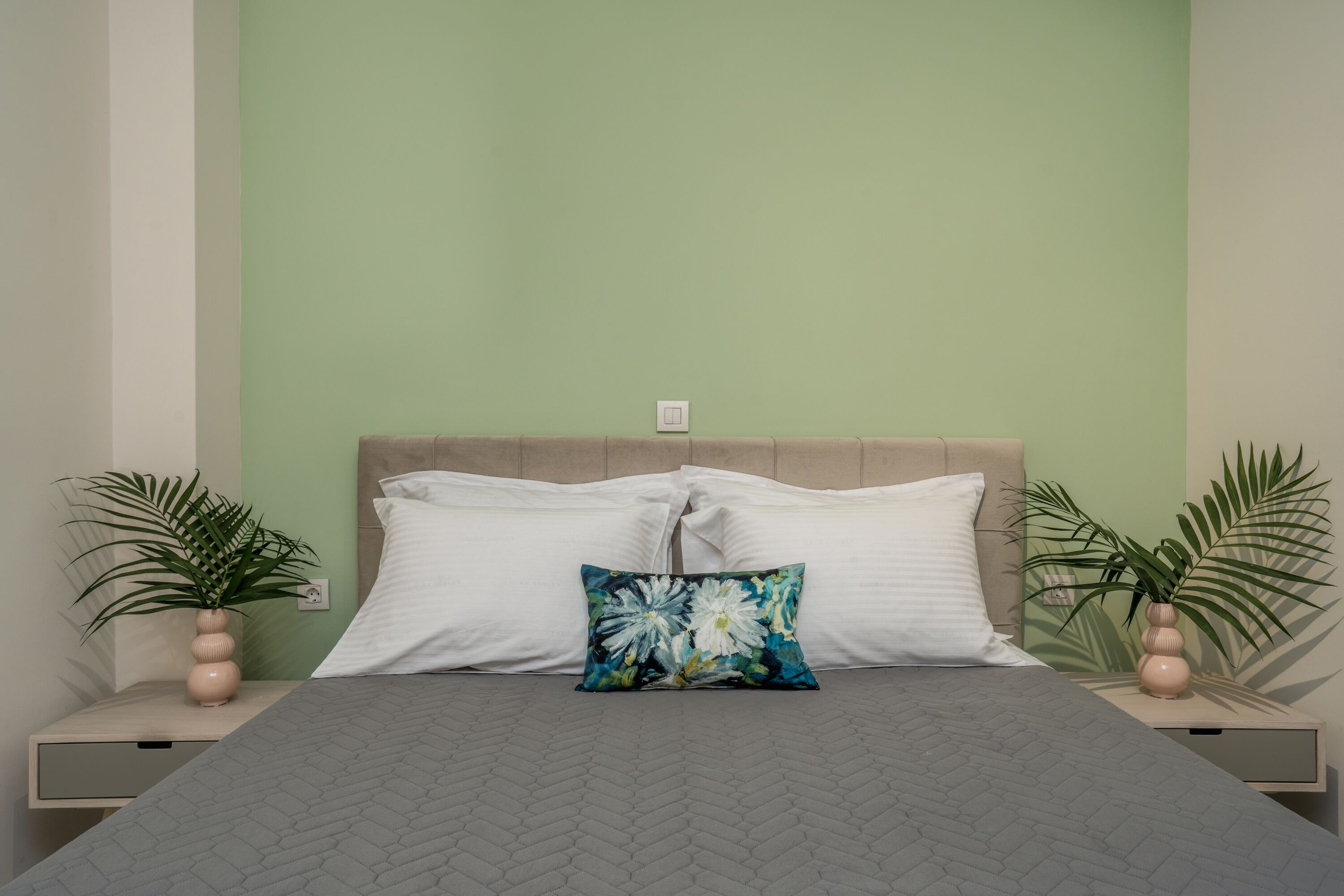 A bed with a grey quilt, two white pillows, and a floral pillow. There are two bedside tables with potted plants.