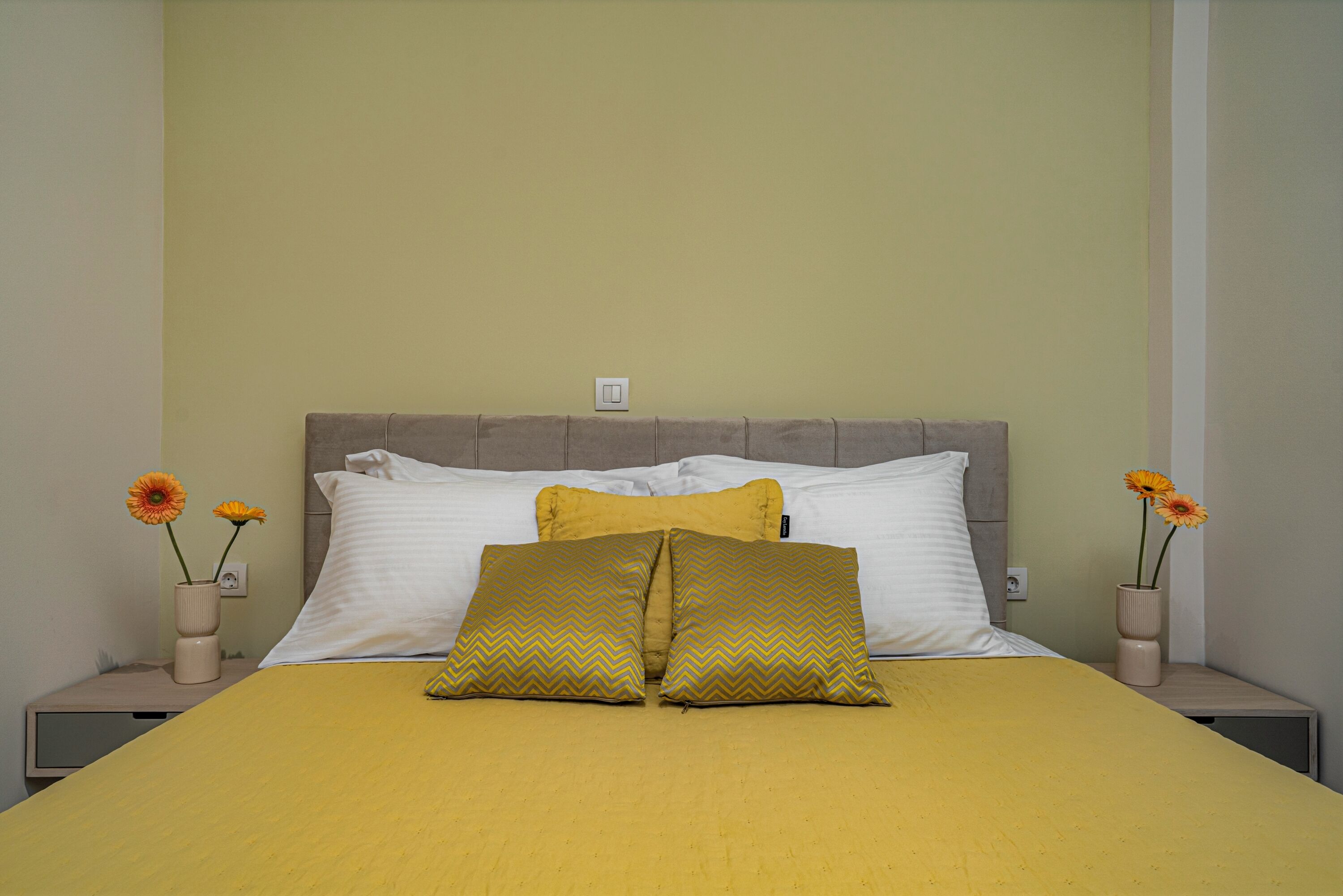 A bed with a yellow bedspread, two white pillows, and two gold patterned pillows. There are two vases with flowers on either side of the bed.