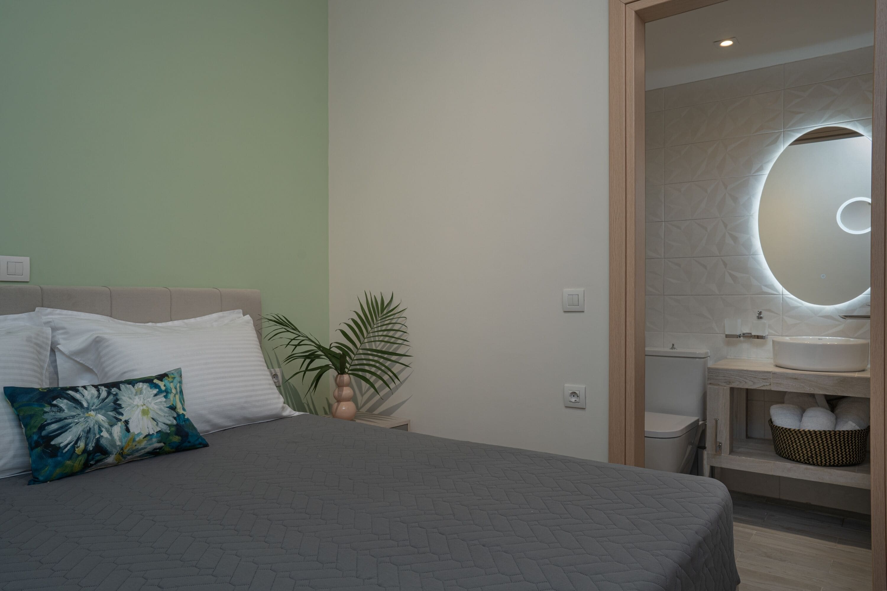 A bedroom with a bed, a bedside plant, and a bathroom with a modern sink and mirror.