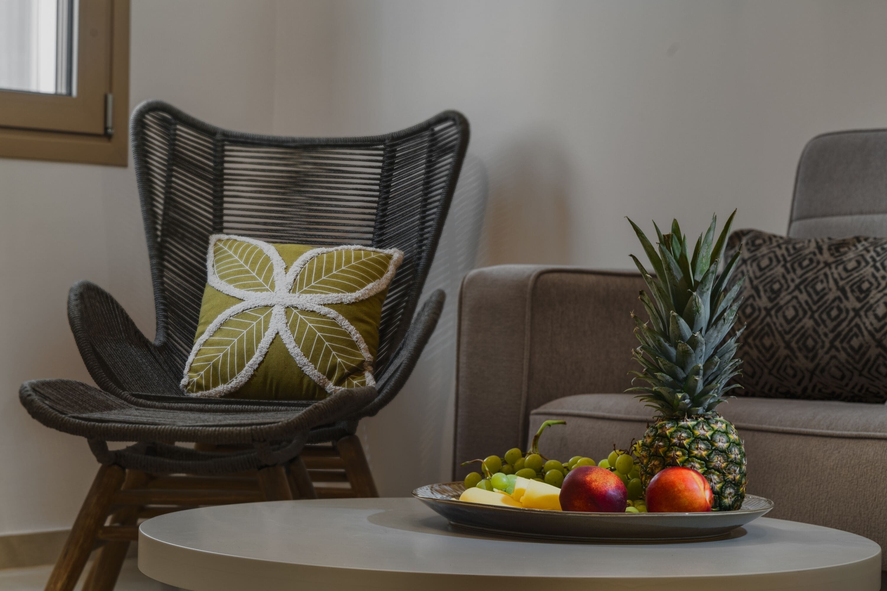 Une chaise en osier avec un coussin orné de motifs de feuilles, un panier de fruits contenant de l’ananas, des raisins et des pommes, et un canapé en arrière-plan.