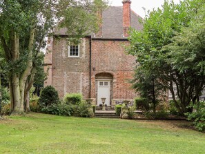 A traditional brick house with a white door and a small window, surrounded by lush greenery and trees.