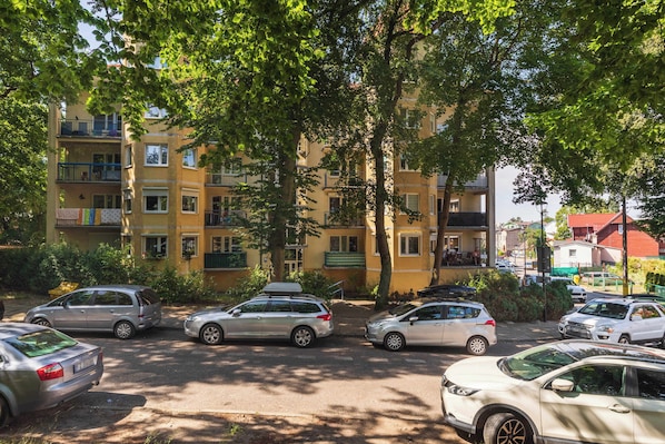 Un parking avec plusieurs voitures, un immeuble jaune doté de balcons et des arbres qui offrent de l’ombre.