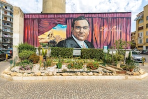 A mural of a man in a suit with a curtain backdrop, surrounded by a landscaped garden with various plants and a cobblestone street.