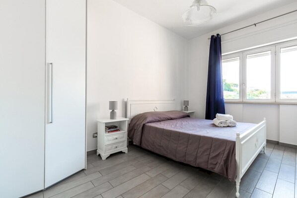 A bedroom with a bed, a nightstand, a window with curtains, and a view of greenery outside.