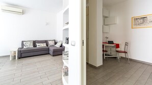 En moderne stue med en grå sofa, et lille bord og en rød stol. Væggen er hvid med et indrammet billede, og loftet har en detaljeret struktur.