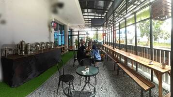 An indoor cafe with tables and chairs, a counter with kitchen equipment, and large windows.