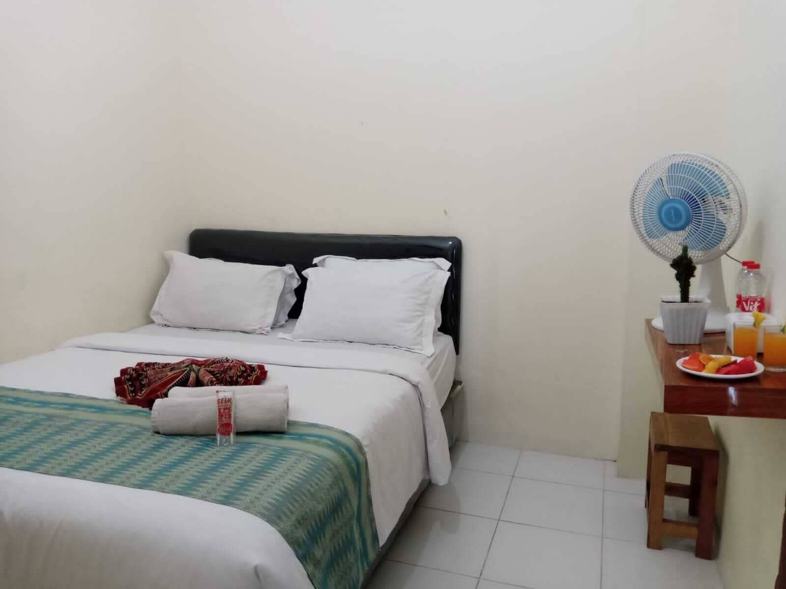Une chambre d’hôtel avec un lit, un bureau équipé d’un ventilateur et une petite table sur laquelle se trouvent une assiette de fruits et une bouteille.