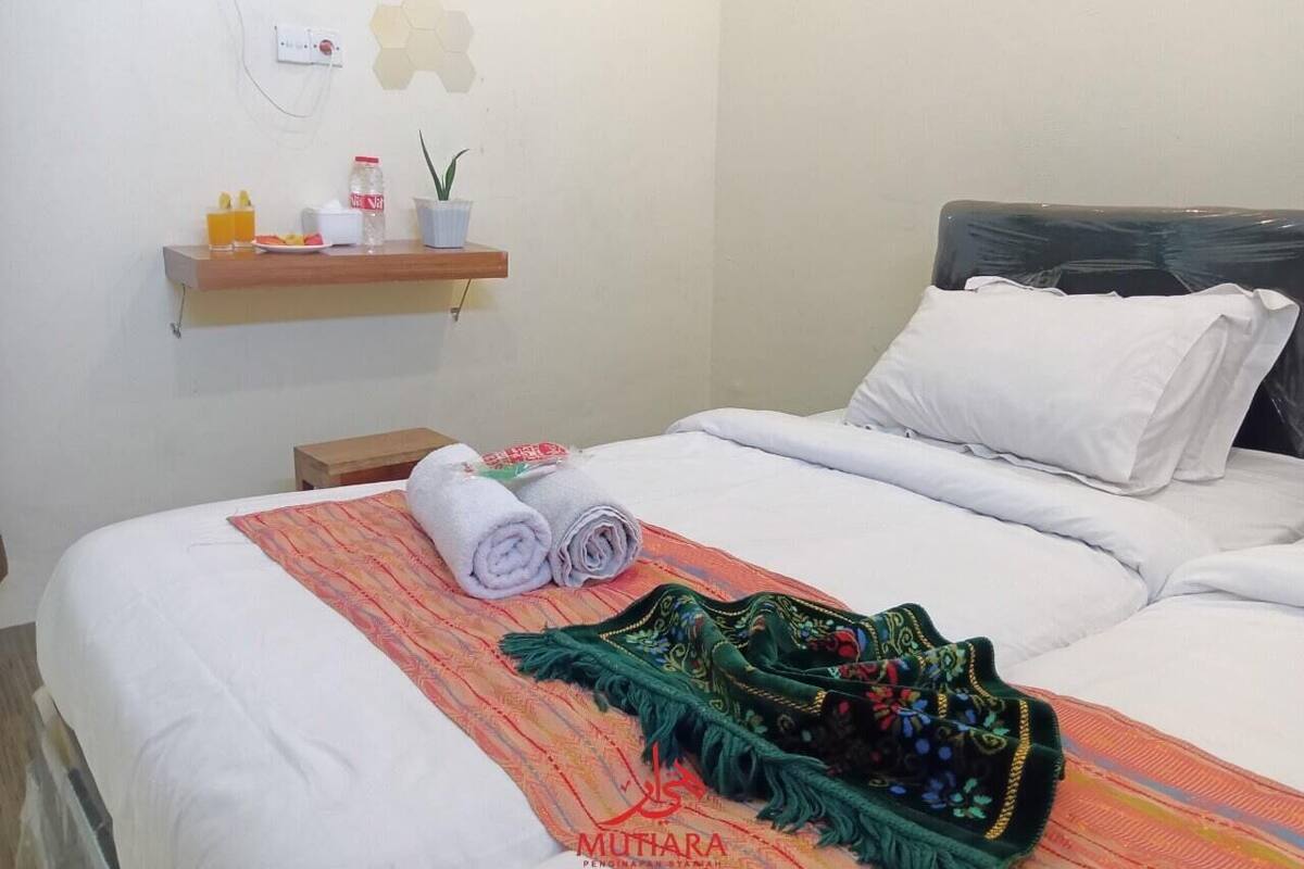 A neatly made bed with white linens, a green decorative throw, and two rolled towels on a wooden shelf above.