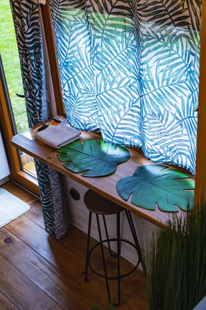 Un escritorio de madera con un cojín grande con forma de hoja, un taburete pequeño y una ventana con cortinas de estampado de hojas tropicales.