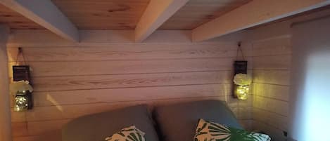 Una habitación acogedora con paredes de madera, una cama con almohadas estampadas con hojas verdes y dos lámparas fijadas a la pared.