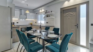 A modern kitchen with white cabinets, a built-in oven, and a dining area with teal chairs and a glass table. - Villa Agalliu (Zakynthos)